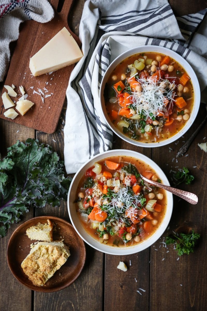 Rustic Minestrone Soup with Rice and Kale in two white bowls on a wooden backdrop with a plate of cornbread to the side.