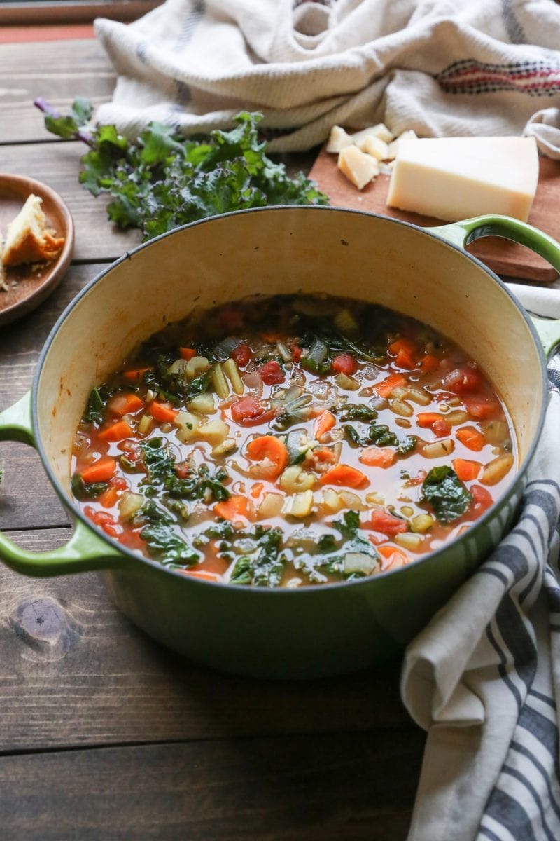 Big Dutch oven full of minestrone soup with a napkin to the side and parmesan cheese on a wooden cutting board.