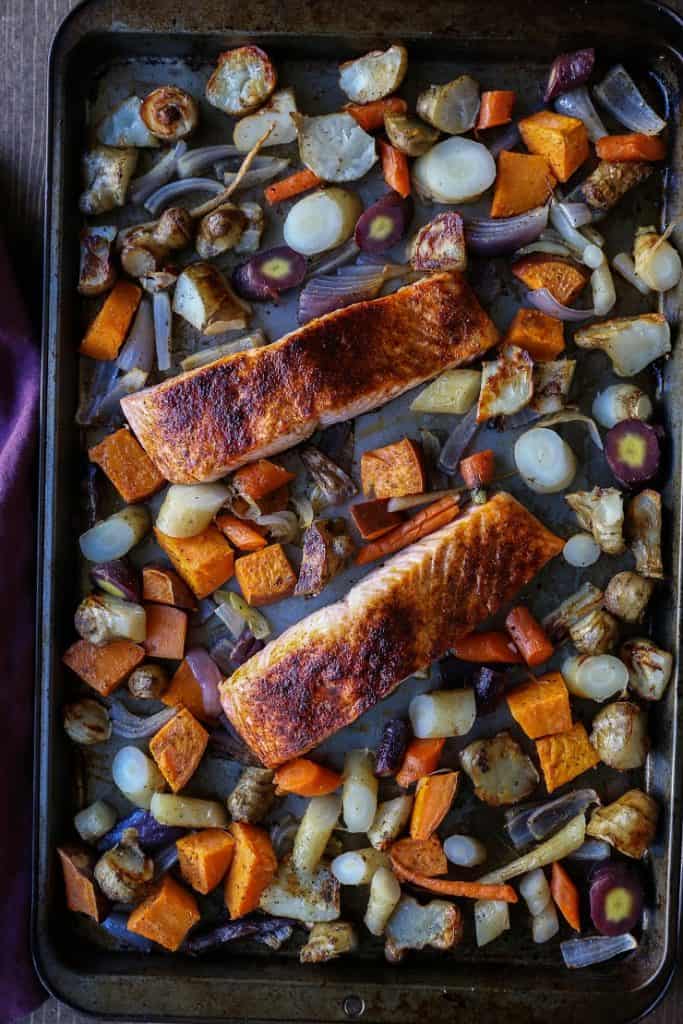 Sheet pan salmon fillets rest on a baking sheet, surrounded by an assortment of roasted vegetables—carrots, sweet potatoes, onions, and parsnips—for a colorful and wholesome meal.