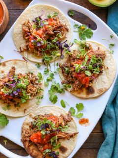 Top down photo of white platter of shredded beef tacos with salsa and microgreens on top of the tacos. Set on a wooden backdrop with a dark blue napkin to the side.