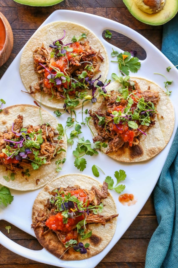 Top down photo of white platter of shredded beef tacos with salsa and microgreens on top of the tacos. Set on a wooden backdrop with a dark blue napkin to the side.