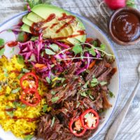 Top down photo of a blue-rimmed bowl with shredded beef, yellow rice, avocado, and slaw inside for the most delicious burrito bowl.