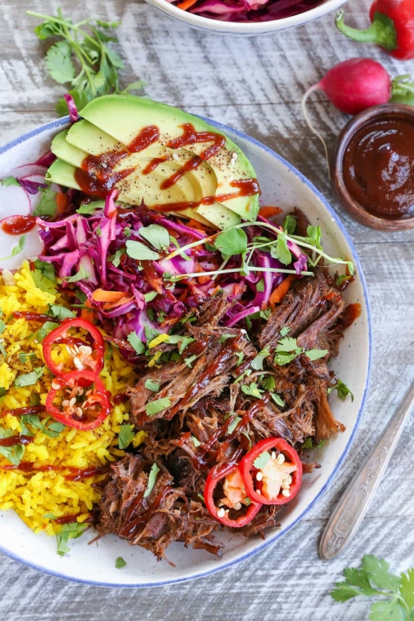 Top down photo of a blue-rimmed bowl with shredded beef, yellow rice, avocado, and slaw inside for the most delicious burrito bowl.