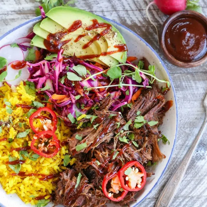 Top down photo of a blue-rimmed bowl with shredded beef, yellow rice, avocado, and slaw inside for the most delicious burrito bowl.