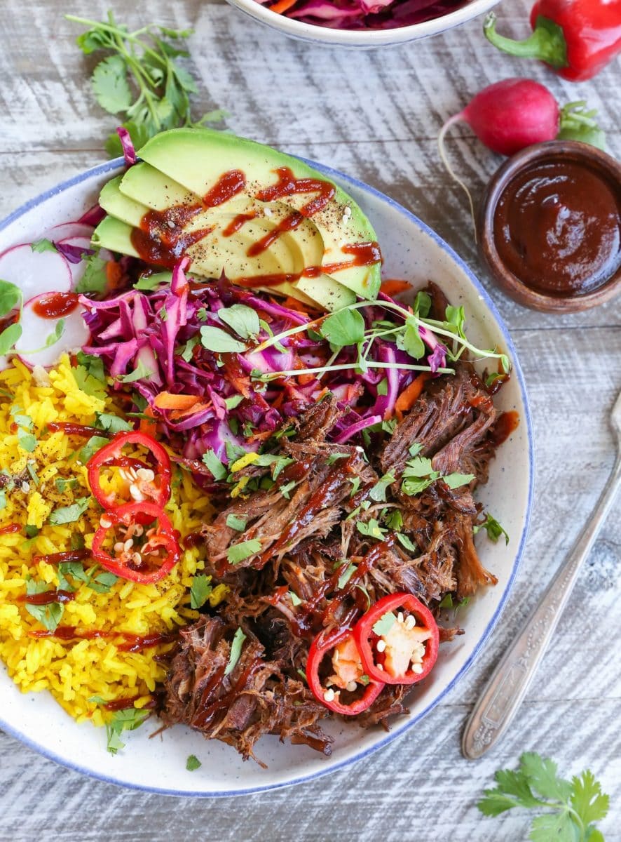 Top down photo of a blue-rimmed bowl with shredded beef, yellow rice, avocado, and slaw inside for the most delicious burrito bowl.