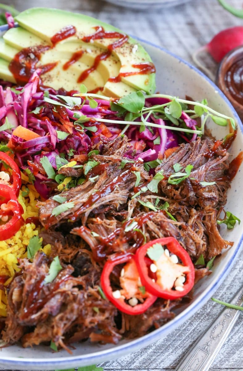 Closeup image of shredded beef inside of a blue-rimmed bowl with rice, avocado, and slaw for a complete meal.