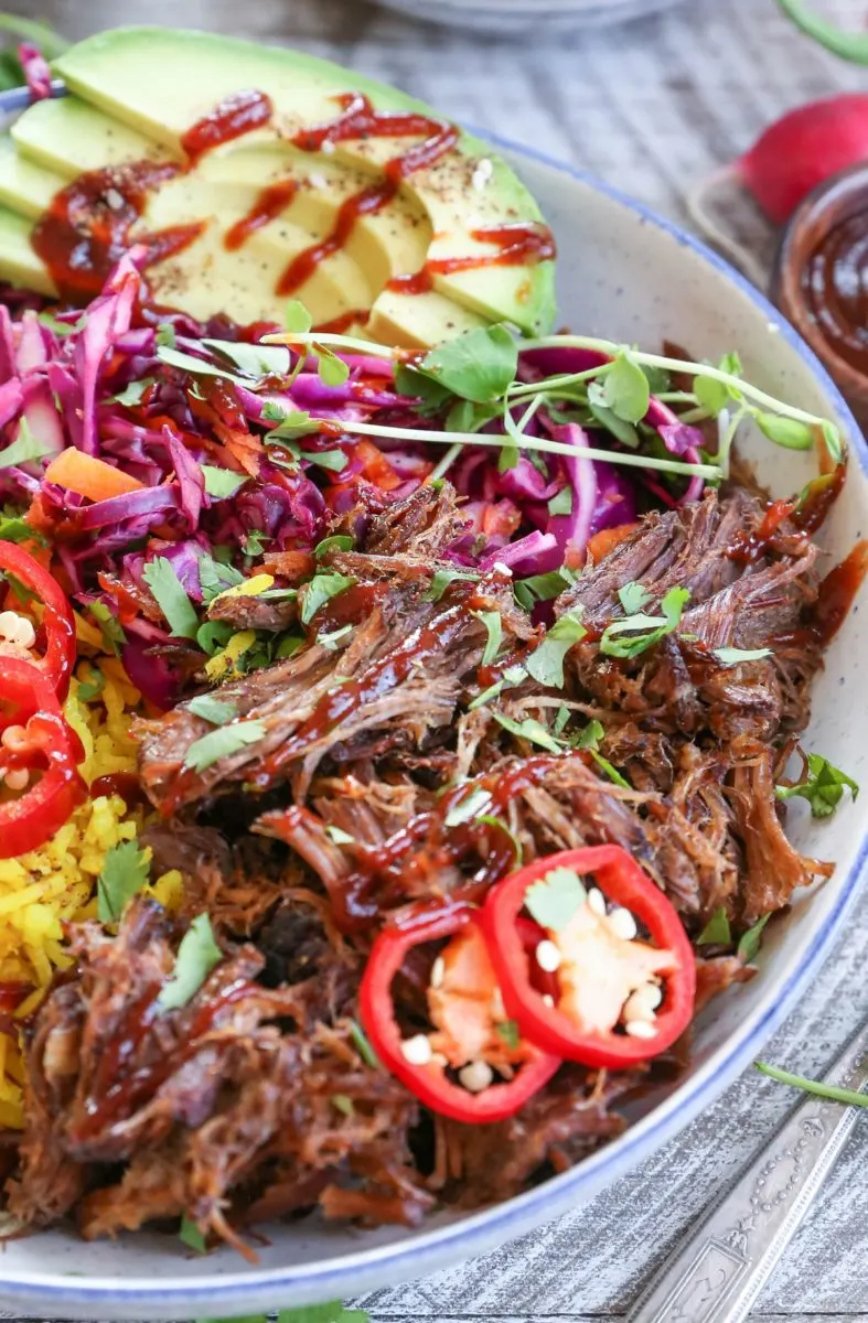 Closeup image of shredded beef inside of a blue-rimmed bowl with rice, avocado, and slaw for a complete meal.
