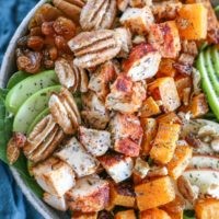 Spinach salad in a big rustic bowl with chicken, butternut squash, raisins, pecans, and apples on top, ready to eat.