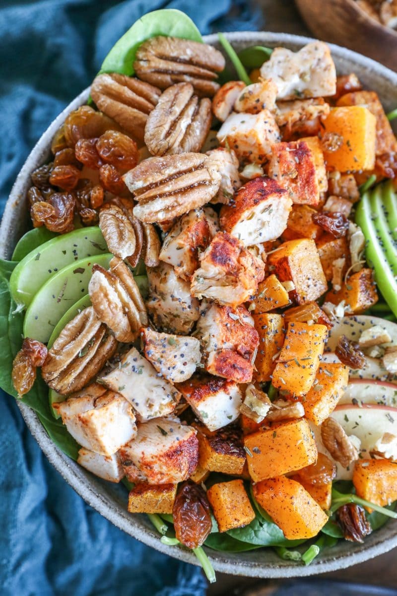 Spinach salad in a big rustic bowl with chicken, butternut squash, raisins, pecans, and apples on top, ready to eat.