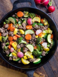 Close up of a cast iron skillet with ground beef and vegetables inside sitting on a wooden backdrop with a golden napkin.