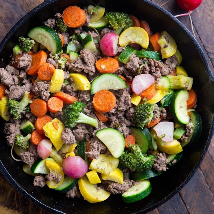 Close up of a cast iron skillet with ground beef and vegetables inside sitting on a wooden backdrop with a golden napkin.