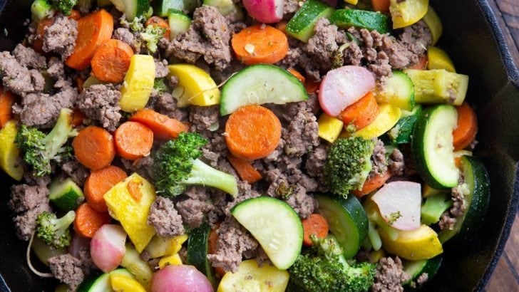 Close up of a cast iron skillet with ground beef and vegetables inside sitting on a wooden backdrop with a golden napkin.