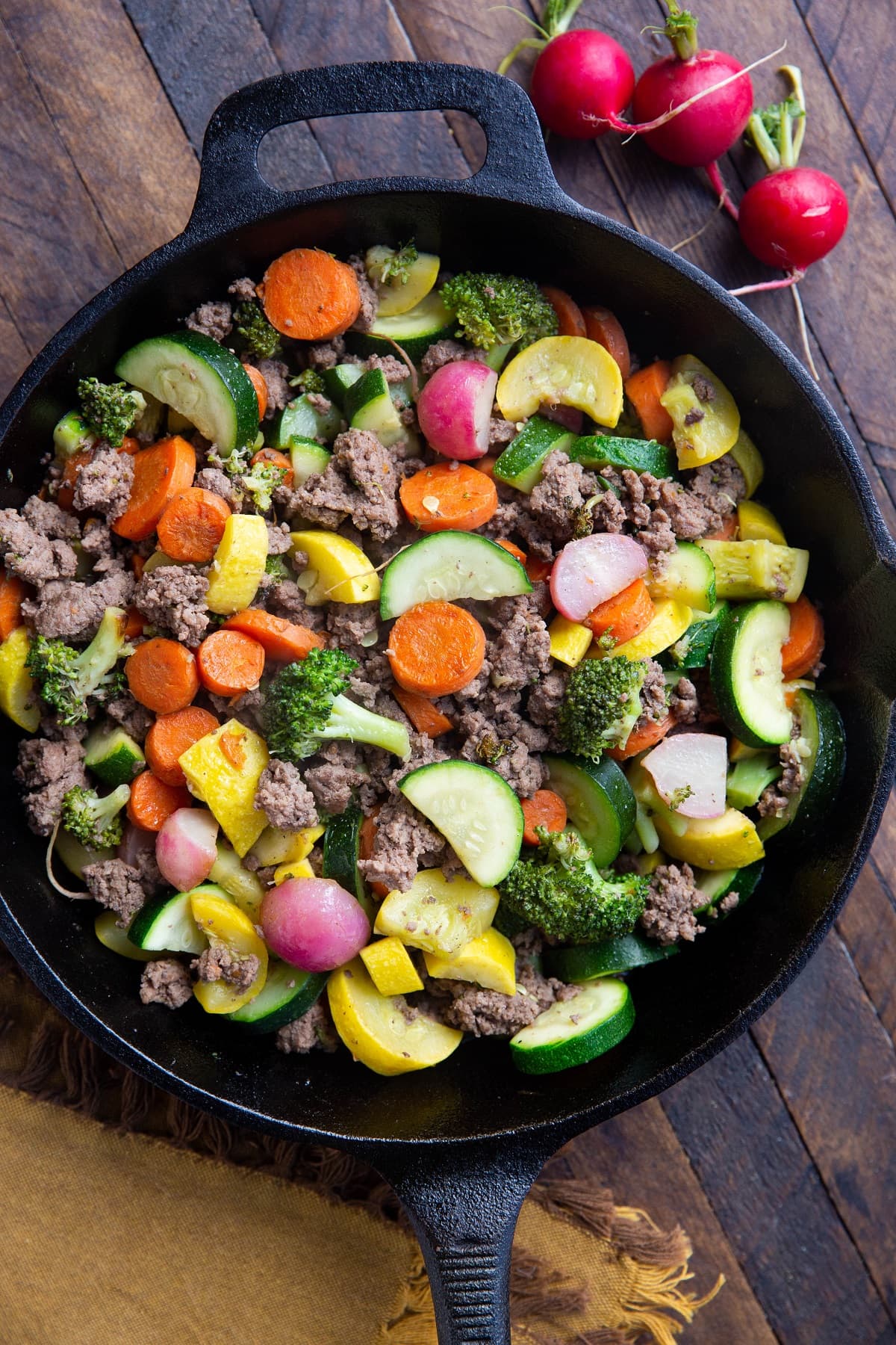 Close up of a cast iron skillet with ground beef and vegetables inside sitting on a wooden backdrop with a golden napkin.