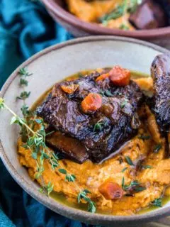Beef short ribs on top of mashed sweet potatoes in a large bowl, ready to eat! A dark blue napkin to the side and a rustic wood backdrop.