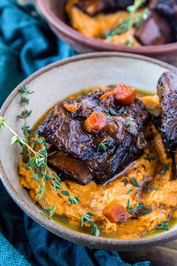 Beef short ribs on top of mashed sweet potatoes in a large bowl, ready to eat! A dark blue napkin to the side and a rustic wood backdrop.