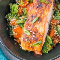 Top down photo of a black bowl of salmon, rice, and vegetables with chop sticks to the side and green onion sprinkled on top of the food with a blue napkin to the side.