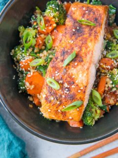 Top down photo of a black bowl of salmon, rice, and vegetables with chop sticks to the side and green onion sprinkled on top of the food with a blue napkin to the side.