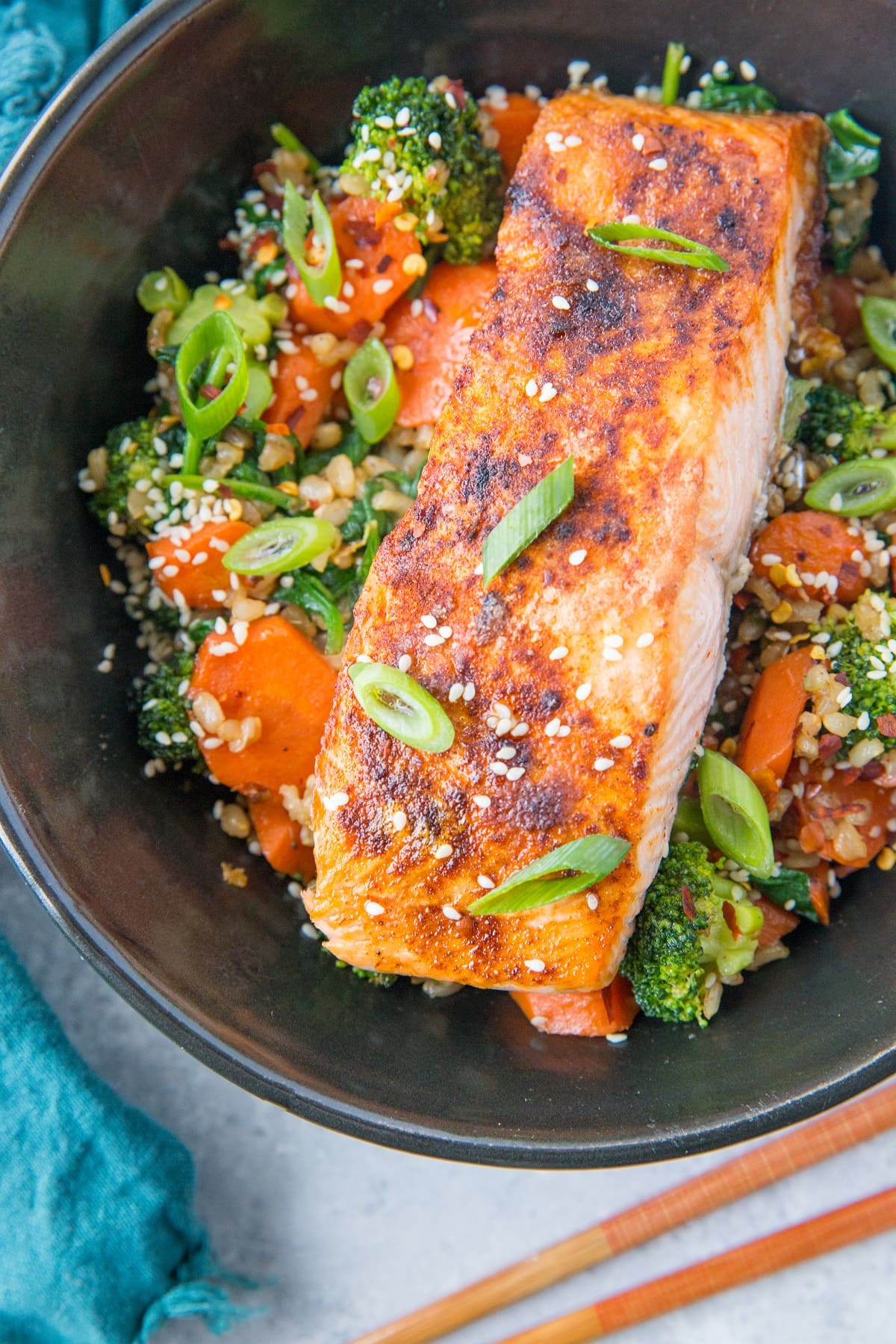 Top down photo of a black bowl of salmon, rice, and vegetables with chop sticks to the side and green onion sprinkled on top of the food with a blue napkin to the side.