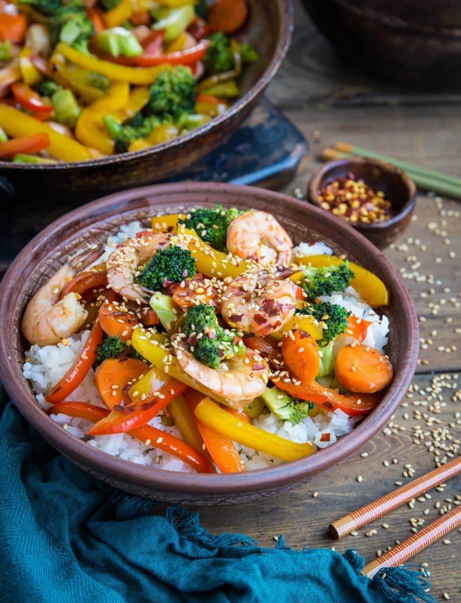 Big bowl of shrimp stir fry with a dark blue napkin to the side with chopsticks and a skillet of stir fry in the background.