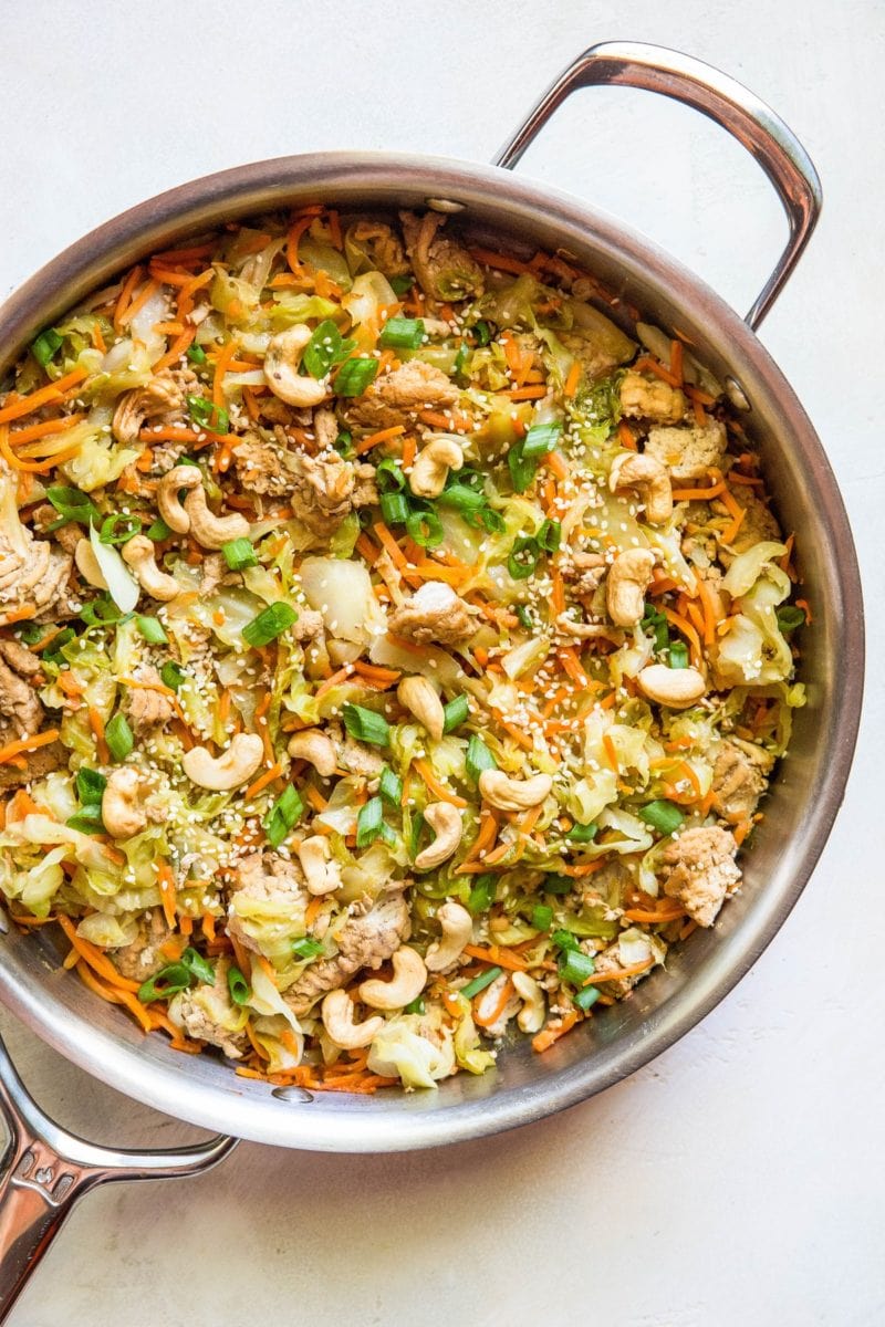 A stainless steel skillet with sauteed cabbage, carrots, ground turkey, and roasted cashews for making egg roll in a bowl