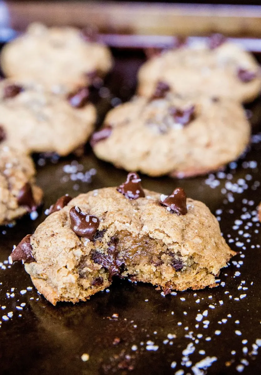 Close up on a chocolate chip cookie sitting on a baking sheet with a bite taken out so you can see the gooey inside.