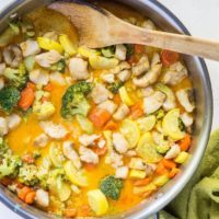 Stainless steel skillet with red curry with fish and vegetables inside and a wooden spoon with a green napkin wrapped around handle of skillet