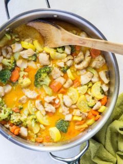 Stainless steel skillet with red curry with fish and vegetables inside and a wooden spoon with a green napkin wrapped around handle of skillet