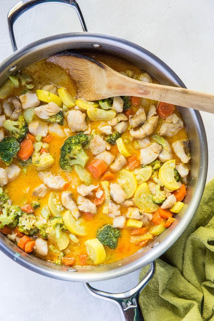Stainless steel skillet with red curry with fish and vegetables inside and a wooden spoon with a green napkin wrapped around handle of skillet