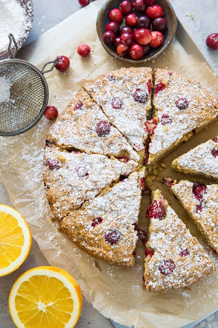 cranberry orange cake on a baking sheet with slices, sprinkled with powdered sugar with fresh cranberries and oranges surrounding cake
