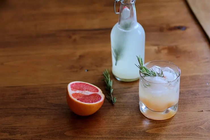 A bottle and a glass of grapefruit rosemary ginger beer sitting on a table with half a grapefruit and sprigs of rosemary