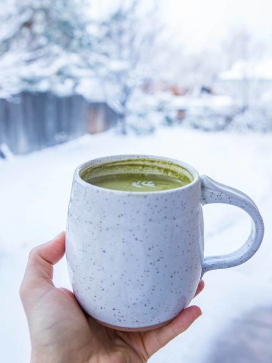 A hand holds a speckled white ceramic mug filled with creamy bulletproof matcha latte, set against a snowy winter landscape with blurred trees in the background.