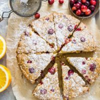 grain-free cranberry orange cake on parchment paper on top of a baking sheet, sprinkled with powdered sugar with fresh cranberries and oranges next to the cake