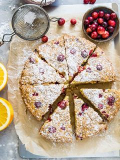 grain-free cranberry orange cake on parchment paper on top of a baking sheet, sprinkled with powdered sugar with fresh cranberries and oranges next to the cake