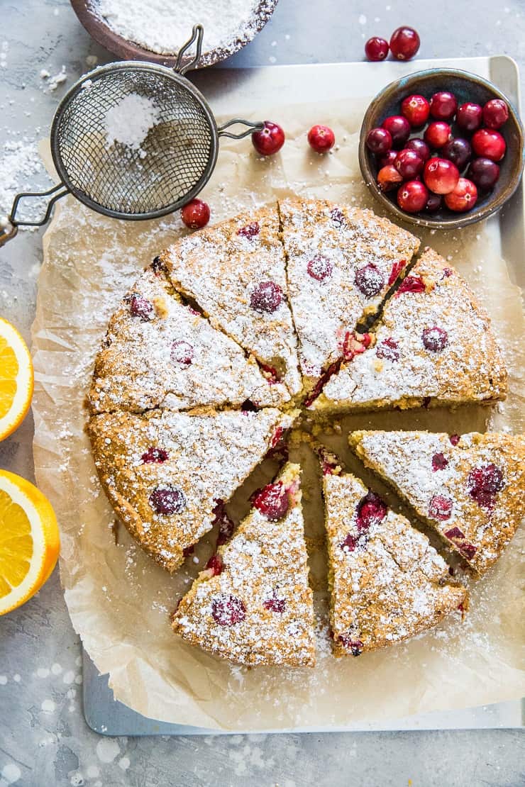 grain-free cranberry orange cake on parchment paper on top of a baking sheet, sprinkled with powdered sugar with fresh cranberries and oranges next to the cake