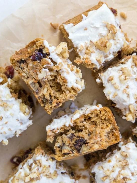 Sliced pieces of low-carb carrot cake topped with white frosting and chopped walnuts, showing a moist, crumbly texture with visible bits of carrot and raisins, arranged on parchment paper.