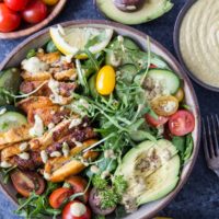 Top down image of whole bowl of crispy chicken salad with avocado, arugula, lemon tahini dressing, and a blue background.