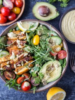 Top down image of whole bowl of crispy chicken salad with avocado, arugula, lemon tahini dressing, and a blue background.