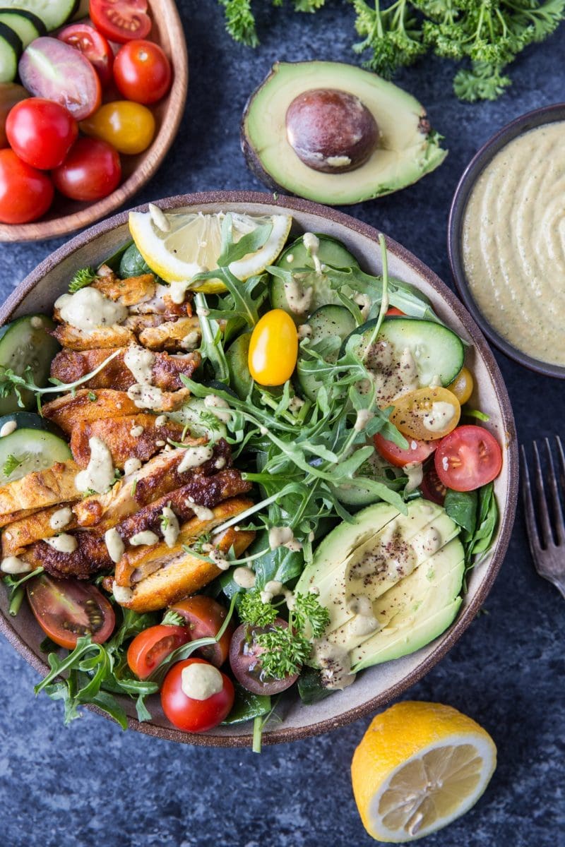 Top down image of whole bowl of crispy chicken salad with avocado, arugula, lemon tahini dressing, and a blue background.