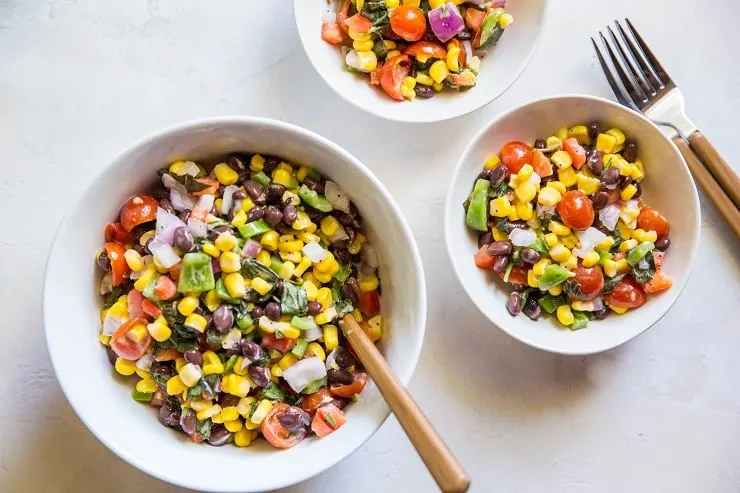 one large bowl and two small bowls of black bean corn salad with two forks on the side on a white backdrop