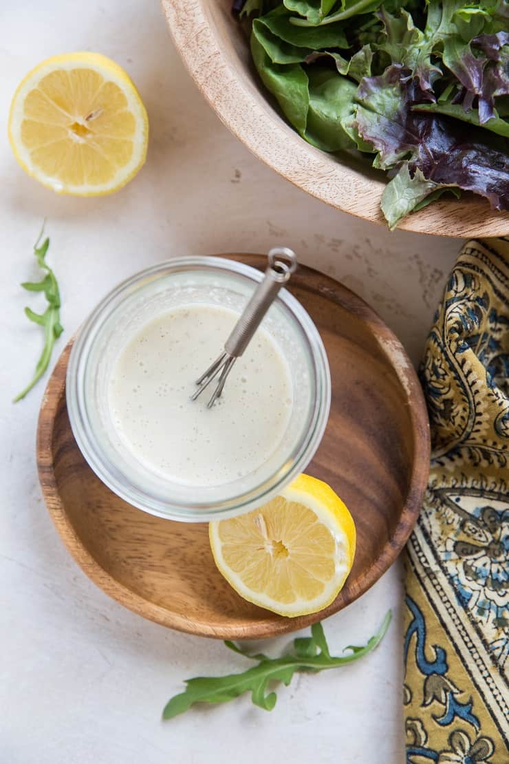 Top down photo of jar of caesar dressing with bowl of lettuce to the side with a napkin and halved lemons