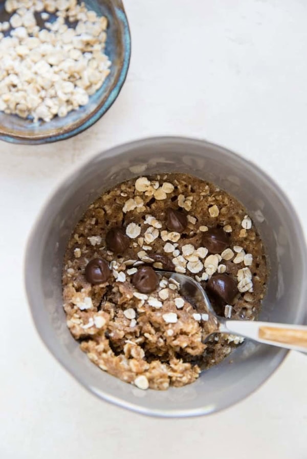 A gray bowl filled with oatmeal, chocolate chips, and a spoon evokes the cozy feel of an oatmeal cookie in a mug. Next to it is a small bowl of dry oats, all set on a light-colored surface.