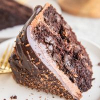 Thick slice of chocolate layer cake with chocolate frosting and chocolate ganache on a white plate with a gold fork.