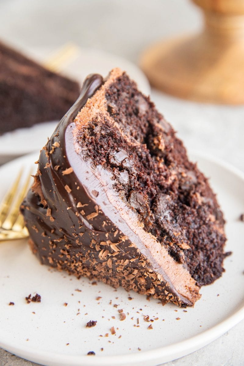 Thick slice of chocolate layer cake with chocolate frosting and chocolate ganache on a white plate with a gold fork.