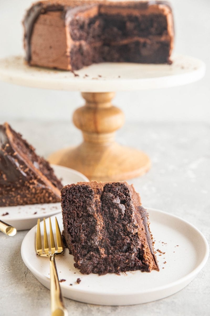 Insanely rich and delicious gluten-free chocolate cake made with no dairy or refined sugar. Two slices sitting on a white plate with a golden fork and the rest of the cake on a cake stand in the background