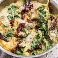 Skillet with spaghetti squash and sun-dried tomatoes, artichoke hearts and spinach with a creamy sauce. Grey napkin to the side and dried herbs all around.