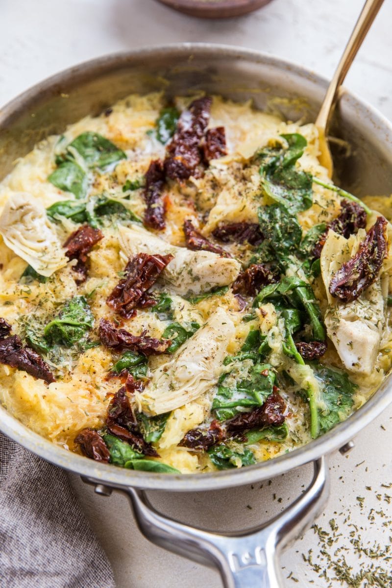 Skillet with spaghetti squash and sun-dried tomatoes, artichoke hearts and spinach with a creamy sauce. Grey napkin to the side and dried herbs all around.