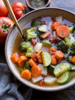 Big ceramic bowl of healthy vegetable soup with a gold spoon, ready to eat. A grey napkin and a fresh tomato to the side.