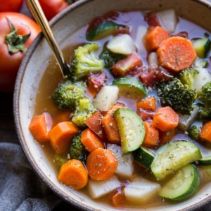 Big ceramic bowl of healthy vegetable soup with a gold spoon, ready to eat. A grey napkin and a fresh tomato to the side.