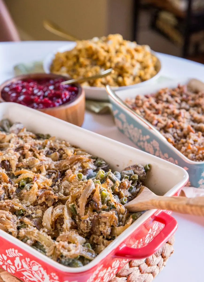 Casserole dish with green bean casserole and other side dishes like stuffing and cranberry sauce in the background.