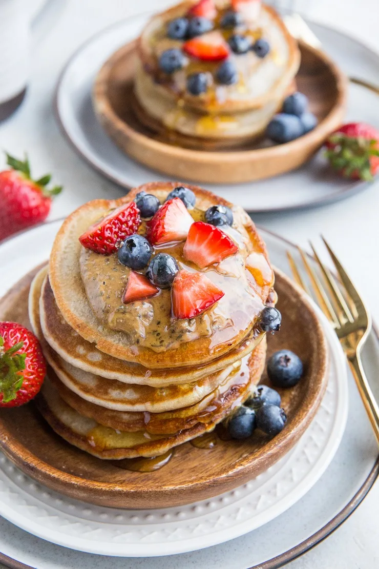 Two plates with stacks of kombucha pancakes with nut butter berries and honey on top